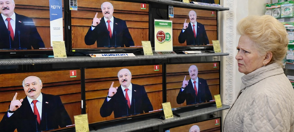 Возрастная женщина с начесом смотрит на ряд телевизоров в магазине бытовой техники; на всех экранах транслируется выступление Александра Лукашенко.
